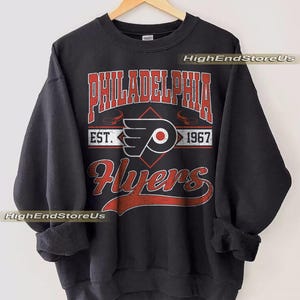 Peut inclure: Sweat-shirt noir avec l'inscription "PHILADELPHIA Flyers" en rouge et blanc. Le design comprend le logo de l'équipe et "EST. 1967". Le sweat-shirt est suspendu à un cintre en bois.