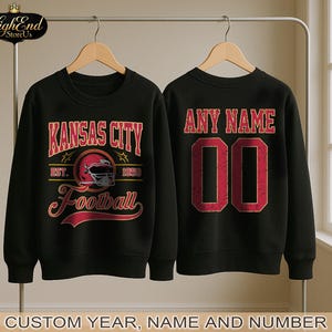 Puede incluir: Dos sudaderas negras con tem&aacute;tica de f&uacute;tbol. Una presenta "Kansas City Football" en letras rojas y doradas con un gr&aacute;fico de casco. La otra tiene "ANY NAME" y el n&uacute;mero "00" en rojo. El texto "CUSTOM YEAR, NAME AND NUMBER" est&aacute; en la parte inferior.