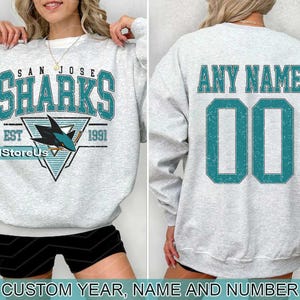 May include: A grey sweatshirt with a San Jose Sharks logo on the front and a blank space on the back for customisation. The logo features a teal shark fin and the text "San Jose Sharks Est 1991". The back of the sweatshirt has the text "Any Name" and a blank space for a number.