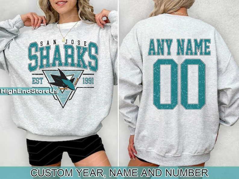 May include: A grey sweatshirt with a San Jose Sharks logo on the front and a blank space on the back for customisation. The logo features a teal shark fin and the text "San Jose Sharks Est 1991". The back of the sweatshirt has the text "Any Name" and a blank space for a number.