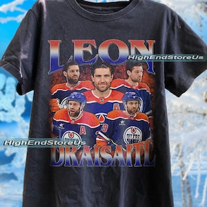 Könnte beinhalten: Schwarzes T-Shirt mit einem Vintage-Grafikdesign von Eishockeyspielern in blauen und orangefarbenen Trikots. Das Shirt trägt den Namen "LEON" in einem Farbverlauf und "DRAISAITL" darunter. Der Text "HighEndStoreUs" ist ebenfalls sichtbar.