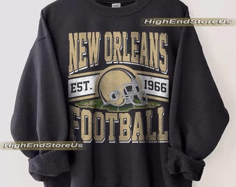 May include: Black sweatshirt featuring a distressed gold and white graphic. The graphic includes the words "NEW ORLEANS FOOTBALL" with a football helmet and the year "1966". The sweatshirt is hanging on a wooden hanger.