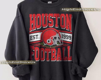 Könnte beinhalten: Schwarzes Sweatshirt mit einem verblichenen rot-weißen Aufdruck "HOUSTON FOOTBALL EST. 1999." Das Design beinhaltet einen roten Footballhelm. Das Sweatshirt hängt an einem Holzkleiderbügel.