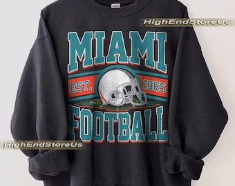 Könnte beinhalten: Schwarzes Sweatshirt mit dem Text "MIAMI FOOTBALL" in türkis-weißen Blockbuchstaben und einer Footballhelm-Grafik. Der Text "EST. 1965" befindet sich über dem Helm. Das Sweatshirt hängt an einem Holzkleiderbügel.