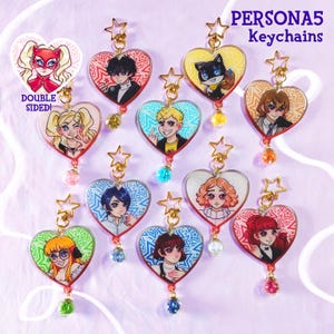 Può includere: Una collezione di portachiavi a forma di cuore con illustrazioni di personaggi in stile anime. Ogni portachiavi è a doppia faccia, con una stella sulla parte superiore e una perla decorativa. Il testo "PERSONA5 Keychains" è visibile.