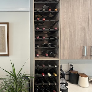 Może przedstawiać: Wysoki, wbudowany drewniany stojak na wino wypełniony różnymi butelkami wina. Stojak jest częścią szafki kuchennej, z rośliną i oprawionym dziełem sztuki widocznym w tle. Butelki wina są w różnych kolorach i rozmiarach.