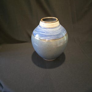 May include: A handcrafted ceramic vase with a rounded body and a narrow neck. The vase features a gradient of blue hues, with a lighter blue band near the top and a darker blue base. The rim is a light beige color with a brown accent.