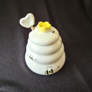 May include: A white ceramic honey pot shaped like a beehive, with a yellow flower on top and small bee illustrations. A heart-shaped dipper rests on the side. The pot has a tiered design and is set against a dark background.