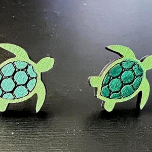 May include: A pair of wooden turtle-shaped earrings. The earrings feature a light green outline with a teal and black patterned shell design. The earrings are small and likely for pierced ears.