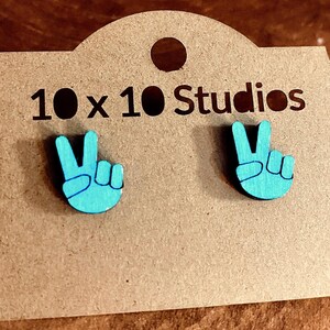 May include: A pair of turquoise peace sign earrings on a brown cardboard display card. The card has the text "10 x 10 Studios" printed in black. The earrings are a vibrant, eye-catching accessory.