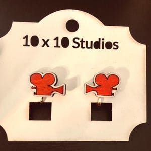 May include: A pair of clip-on earrings featuring a red vintage movie camera design. The earrings are attached to a white card with the text "10 x 10 Studios" printed in black. The earrings have gold-colored clips.