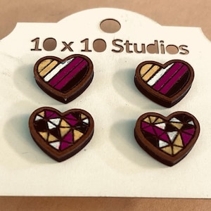 Könnte beinhalten: Vier herzförmige Holzohrringe mit geometrischen Designs. Zwei Ohrringe haben ein Design mit gold, weißen und magenta Streifen. Die anderen beiden haben ein Mosaikmuster in Gold, Weiß und Magenta. Die Ohrringe befinden sich auf einer weißen Karte mit dem Text "10 x 10 Studios".