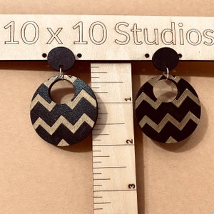 Puede incluir: Pendientes de madera con un diseño de chevron negro y dorado. Los pendientes son redondos con un pequeño círculo en la parte superior y un gancho plateado. El texto "10 x 10 Studios" es visible en el fondo.