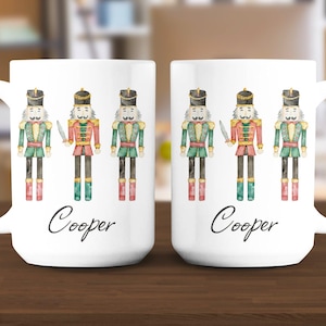 May include: Two white ceramic mugs with a watercolor design of nutcracker soldiers. Each mug features three nutcrackers in red, green, and gold uniforms, with the name "Cooper" printed in black script below the soldiers.