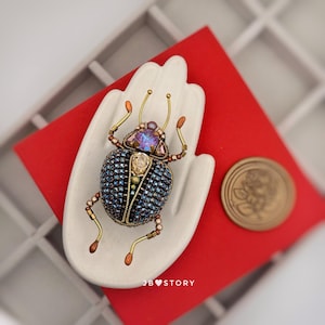 Könnte beinhalten: Ein mit Juwelen besetzter Käfer-Broche mit einem blau-perlenbesetzten Körper, goldfarbenen Beinen und Fühlern sowie einem Kopf aus lila und klarem Kristall. Die Brosche wird auf einem weißen, handförmigen Halter vor rotem Hintergrund präsentiert. Der Text "JB STORY" ist sichtbar.