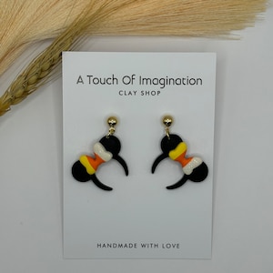 May include: A pair of black and orange Halloween-themed earrings with candy corn designs. The earrings are shaped like Mickey Mouse ears and are handmade with love. The earrings are on a white card with the text "A Touch of Imagination Clay Shop" and "Handmade with Love".