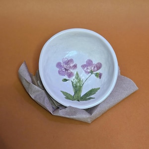 Può includere: Una piccola ciotola in ceramica bianco sporco con un motivo floreale. Due fiori viola con steli e foglie verdi sono dipinti all'interno della ciotola. La ciotola poggia su un pezzo di carta marrone su uno sfondo arancione.