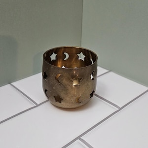May include: A small, cylindrical candle holder made of brass. The holder features cut-out designs of stars and crescent moons. The candle holder is sitting on a white tiled surface.