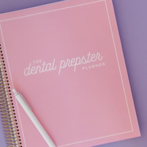May include: A pink spiral-bound planner with the text "dental prepster PLANNER" in white script. A white pen rests diagonally across the planner. The background is a solid purple color.