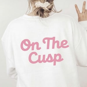Peut inclure: Sweat-shirt blanc avec le texte rose "On The Cusp" dans le dos. Le sweat-shirt est à manches longues et a une coupe décontractée. L'image montre le dos du sweat-shirt.