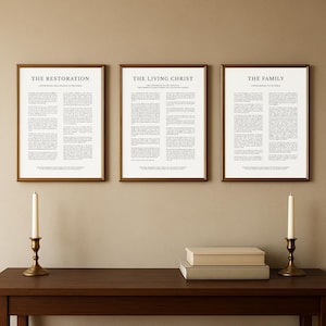 Puede incluir: Tres impresiones enmarcadas con marcos dorados, cada una con texto en fuente negra sobre fondo blanco. Las impresiones se titulan "The Restoration", "The Living Christ" y "The Family". Dos candelabros de latón con velas blancas y dos pilas de libros están sobre una mesa marrón oscuro.