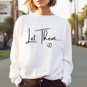 May include: White long-sleeve sweatshirt with the text "Let Them..." and a peace sign.