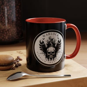 May include: Black coffee mug with a red rim and a white circle graphic featuring a skull with wings and the text "Warlock Eldritch Invocations".