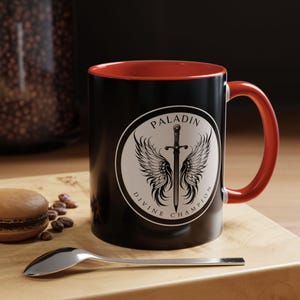 May include: Black coffee mug with a red interior and handle. The mug features a white circular emblem with a sword and wings, and the words "PALADIN DIVINE CHAMPION". A spoon and a pastry are on a wooden surface.