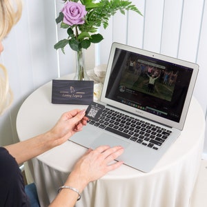 May include: A laptop sits on a round table covered with a white tablecloth. A person holds a card with a QR code. A vase of purple roses and greenery is on the table. The laptop screen displays a video.