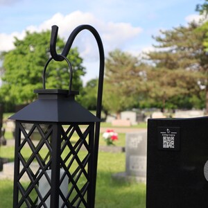 May include: Black metal lantern with a diamond lattice design, hanging from a curved hook. A white candle is visible inside. The lantern is set in a grassy area with a cemetery in the background.