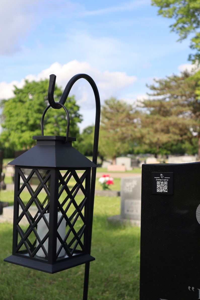 May include: Black metal lantern with a diamond lattice design, hanging from a curved hook. A white candle is visible inside. The lantern is set in a grassy area with a cemetery in the background.