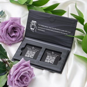 May include: An open black box with two rectangular cards featuring QR codes and the text "Living Legacy". The box is on a white satin surface, with purple roses and green leaves. The box has instructions to scan the Medallion code.