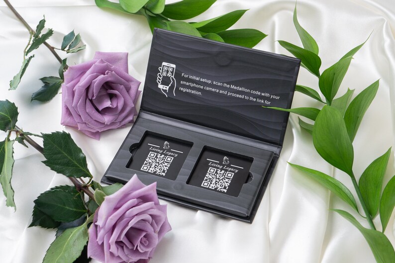 May include: An open black box with two rectangular cards featuring QR codes and the text "Living Legacy". The box is on a white satin surface, with purple roses and green leaves. The box has instructions to scan the Medallion code.