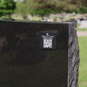 May include: A black granite headstone with a rough-hewn edge. A small black square with the words "Living Legacy" and a QR code is affixed to the headstone. The background shows a cemetery with trees and grass.