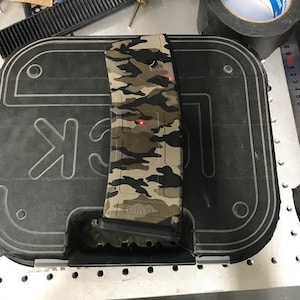 May include: A camouflage-patterned magazine is placed on top of a black case. The magazine features a mix of green, brown, and tan colors. The case has the letters "GLOCK" printed on it. A roll of black tape is visible in the background.