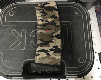 May include: A camouflage-patterned magazine is placed on top of a black case. The magazine features a mix of green, brown, and tan colors. The case has the letters "GLOCK" printed on it. A roll of black tape is visible in the background.