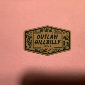 以下が含まれることがあります： 緑色の縁取りと白い内側を持つ茶色の六角形のワッペン。ワッペンには緑色の文字で「OUTLAW HILLBILLY 2024」と印刷されています。