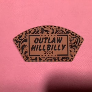 May include: A brown leather patch with a black floral design and the text "OUTLAW HILLBILLY 2024" in a black rectangle.