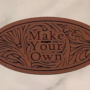 Könnte beinhalten: Ein braunes, ovales Holzschild mit einem dekorativen Blumenmuster und dem Text "Make Your Own" in Schwarz.