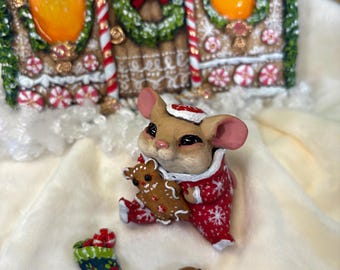 May include: A whimsical Christmas scene featuring a mouse figurine in a red and white snowflake-patterned outfit, holding a gingerbread man. A decorated stocking and a chocolate chip cookie are nearby. The background includes a gingerbread house.