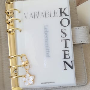 Könnte beinhalten: Ein weißer Plastikordner mit goldenen Ringen und einem weißen Blumenanhänger. Der Ordner hat den Text "VARIABLE KOSTEN" und "Lebensmittel" aufgedruckt. Der Text "MoreLifeDreams" ist unten auf dem Ordner aufgedruckt.
