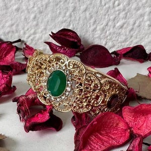 Può includere: Un bracciale dorato con filigrana e una gemma verde ovale al centro, circondata da pietre trasparenti più piccole. Il bracciale è posto su uno sfondo di petali rossi secchi, creando un'estetica lussuosa e ornata.