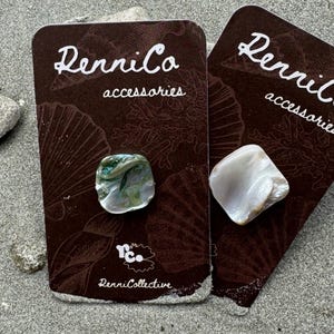 May include: Two square-shaped shell accessories on brown cards with the text "RenniCo accessories". One accessory is white, the other is green and white. The cards are on a sandy surface.