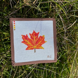 May include: A brown framed canvas with a red and yellow maple leaf design. The canvas is on a bed of green grass.