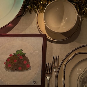 May include: A white needlepoint design with a red and green Christmas pudding with white polka dots. The design is framed in a dark red frame with the initials "RC" in the bottom right corner.