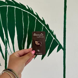 May include: A brown needle minder with the text "RenniCo needle minder" on a brown card. The needle minder has a floral design with the letters "R.C." in the center. The background is a white wall with a green leaf design.