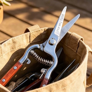 May include: A tan canvas storage bag containing metal garden shears with wooden handles and the engraved text Harvey Scott. The shears feature a spring mechanism and are positioned inside the open-top bag, which has two side handles for carrying.