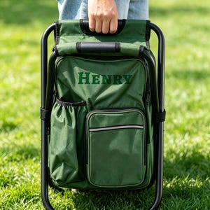 May include: A dark green folding chair with an integrated backpack featuring the name HENRY embroidered in dark green letters. The chair has a black metal frame, a side pocket, a front zippered compartment, and a top handle for carrying.