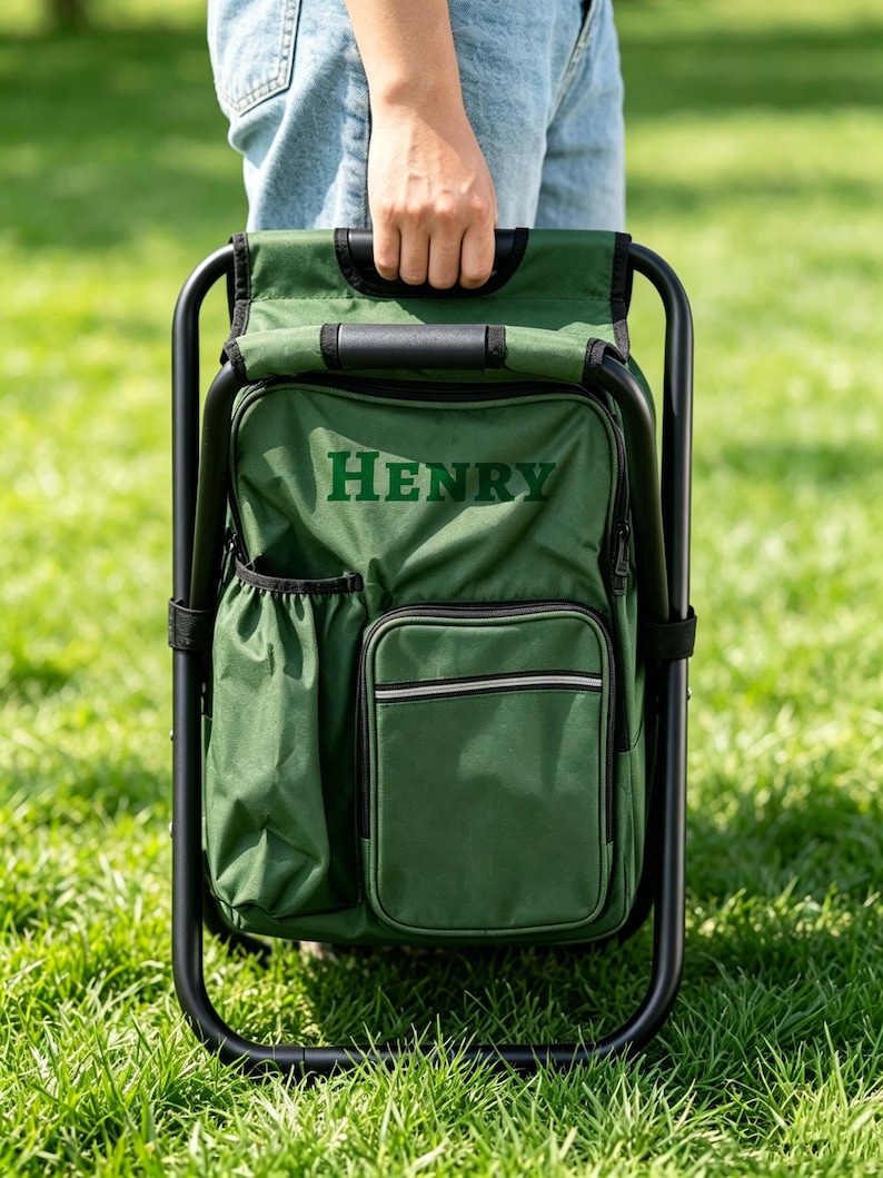 May include: A dark green folding chair with an integrated backpack featuring the name HENRY embroidered in dark green letters. The chair has a black metal frame, a side pocket, a front zippered compartment, and a top handle for carrying.