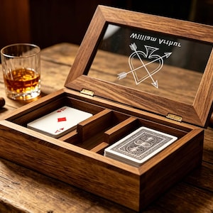 Puede incluir: Una caja rectangular de madera con tapa de cristal abatible que presenta el texto grabado William Arthur y un diseño de corazón y flechas. El interior contiene dos compartimentos con barajas de cartas. La caja es de madera marrón oscura.